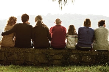 Six people sit side by side on a stone wall, viewed from behind, overlooking a tree-filled landscape. 